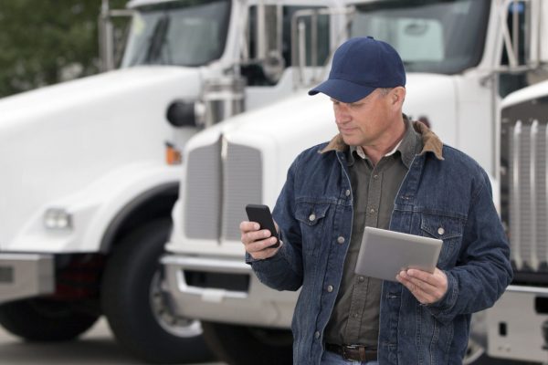 A royalty free image from the trucking industry of a truck driver using a cellphone and tablet computer in front of semi trucks